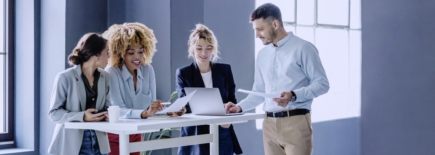 Drei Frauen und ein Mann stehen an einem Stehtisch und schauen auf einen Laptop.