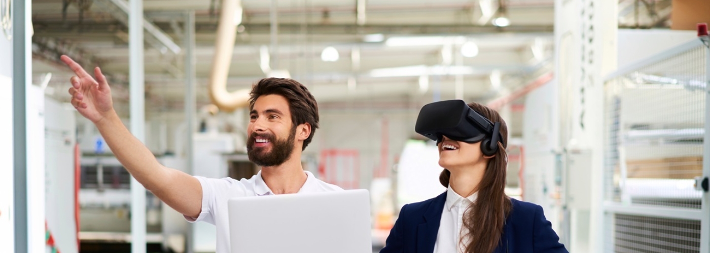 Ein Man steht mit einem Laptop in einem Lager, eine Frau mit einer Virtual-Reality-Brille steht neben ihm.