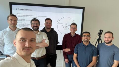 Sieben Männer stehen nebeneinander und schauen in die Kamera, im Hintergrund ist ein Monitor mit einer Grafik zu sehen.