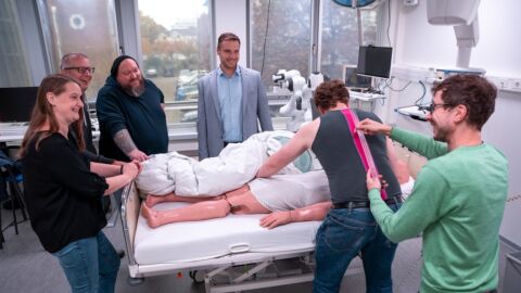 Eine kleine Gruppe von Menschen steht an einem Pflegebett, in dem eine Puppe liegt. Ein Mann dreht diese Puppe, während ein anderer Mann einen Sensorstreifen an seinen Rücken hält.