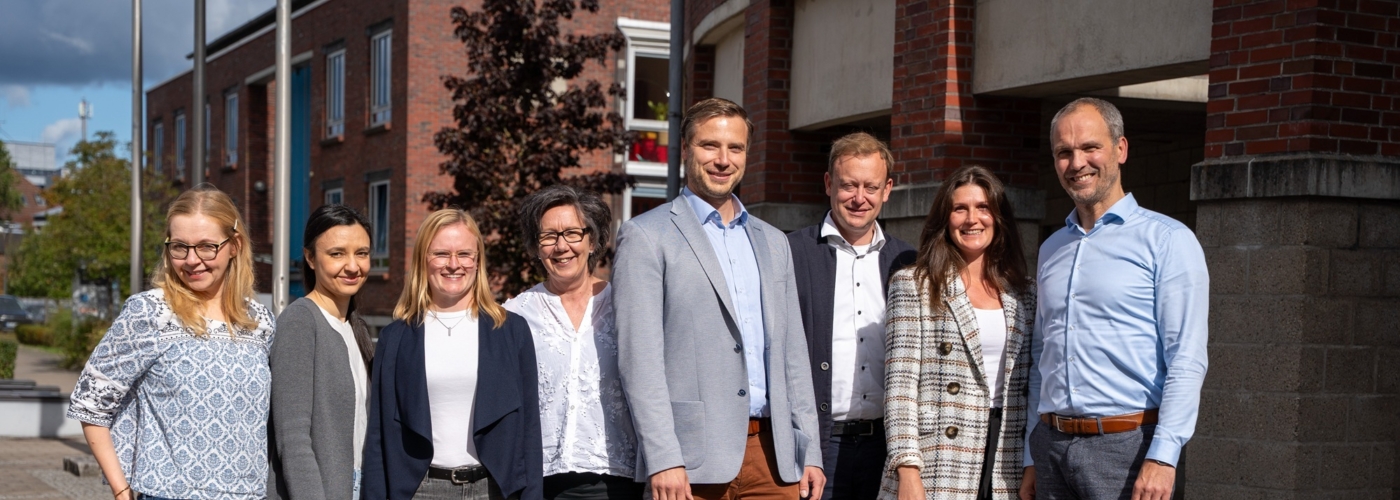 Acht Personen stehen nebeneinander vor einem Bürogebäude und lächeln in die Kamera.