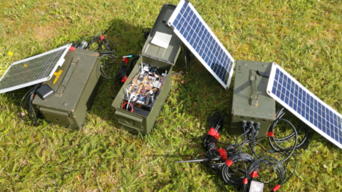 Auf dem Bild sind grüne Metallkisten zusehen, die mit Kabeln und Technik gefüllt sind. Außerdem sind Solarpanele zusehen.