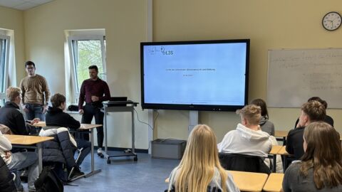 Zwei Männer halten in einem Klassenraum eine Präsentation. Schüler hören zu.