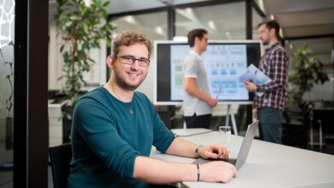 Ein Mann sitzt an einem Tisch mit geöffnetem Laptop und lächelt in die Kamera. Im Hintergrund stehen zwei Männer voreinander und unterhalten sich.