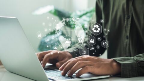 Über Händen auf einer Laptoptastatur schwebt eine Erde mit Symbolen zur Nachhaltigkeit.