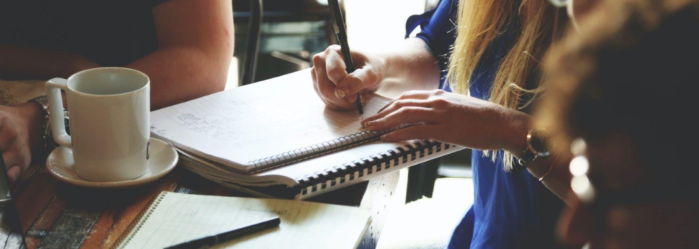 Personen arbeiten an einem Tisch mit Schreibblock und Laptop