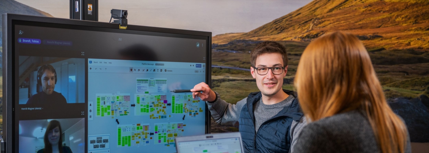 Zwei Personen im Raum bedienen Laptop und großen Bildschirm