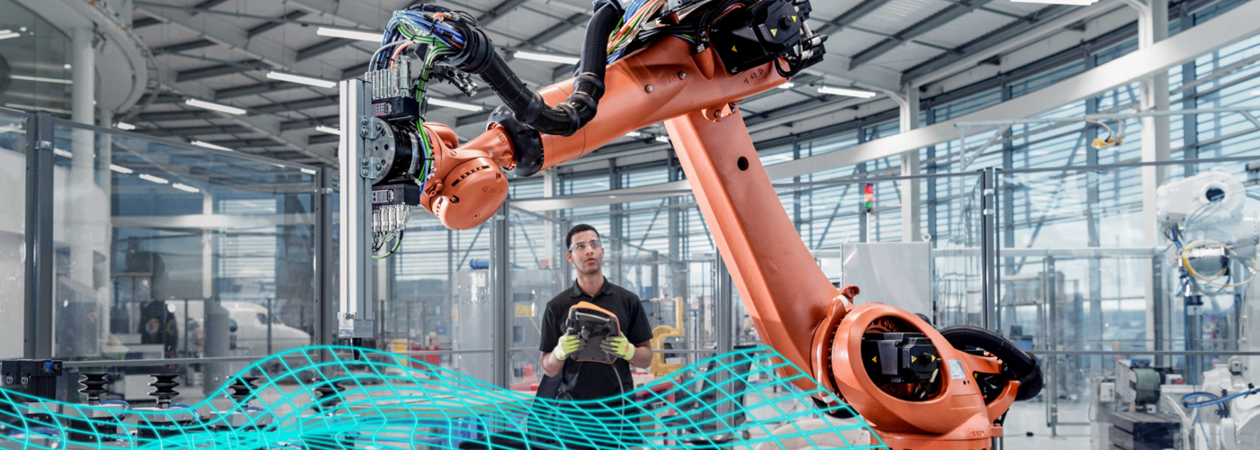 Ein Mann steht mit einer Steuerung in einer Produktionshalle vor einer Maschine mit einem Roboterarm.