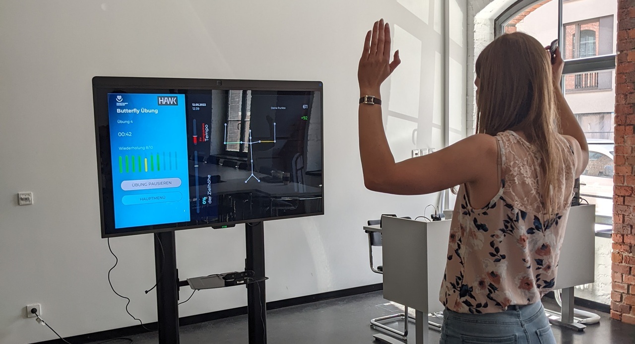 Eine Frau steht vor einem Monitor, auf dem eine Physiotherapie-Übung zu sehen ist. Sie führt die Übung aus.